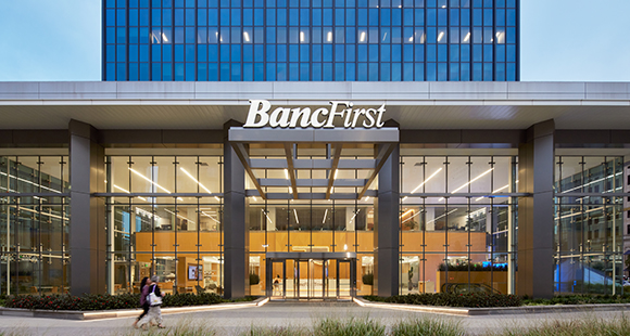 A large glass building with a sign that reads "BancFirst" above the entrance. Outside is a little dark and the interior of the building is lit. A person walks by the entrance.
