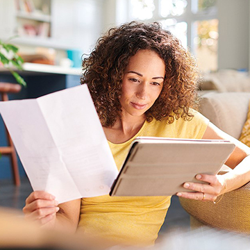 Woman with bill in one hand while looking at tablet in other hand